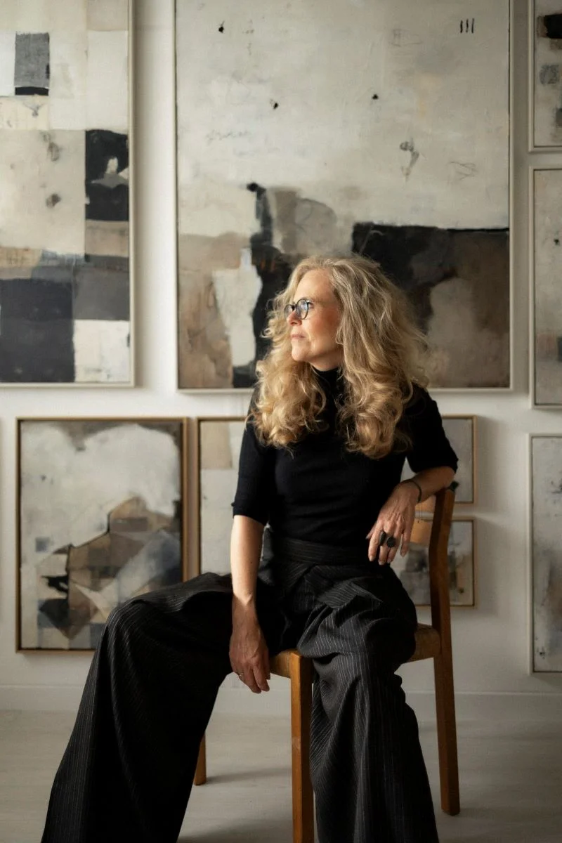 the artist Jolanda wassenaar in here studio, sitting in front of her artwall. sitting on a danish wooden chair in black clothes