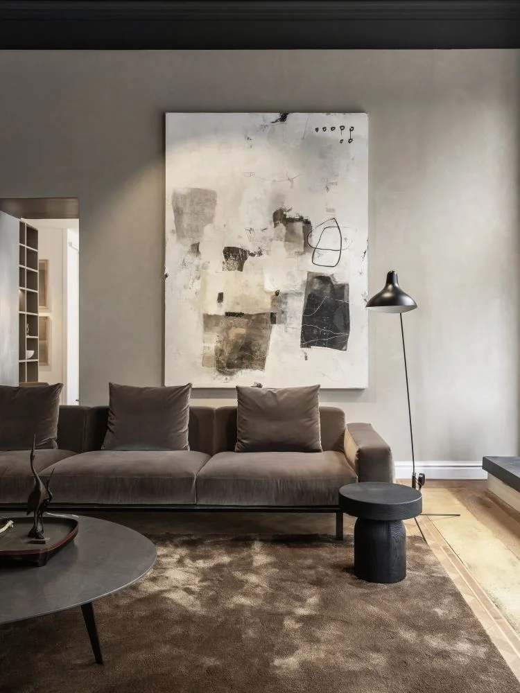 chique interior with a velver brown sofa and a hudge carpet with behind the sofa on the wall a hudge paiting in size 170x230 cm . the colour of the wall is taupe, thats really a good combination and contrast with the light painting. the painting is abstract and mostly white with interesting layers and some marks in black and brown colours.